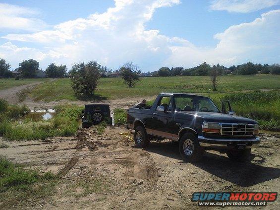 Black Ford Bronco parked on dirt road, driver's door open. Another SUV is stuck in mud in the background