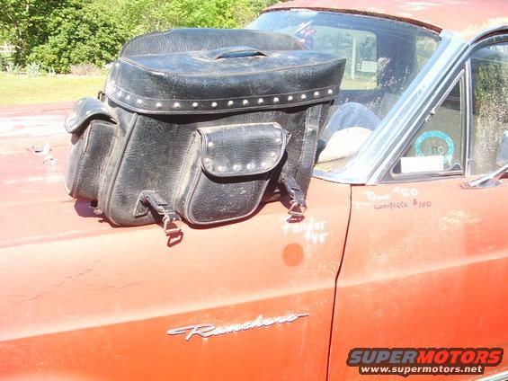Black studded motorcycle saddlebag on the hood of a red car with 'Ranchero' badge. Writing on hood says 'Complete $100'