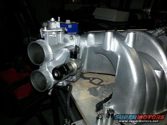 Close-up of a silver engine intake manifold and throttle body assembly, possibly for a Ford vehicle. A blue can is visible in the background