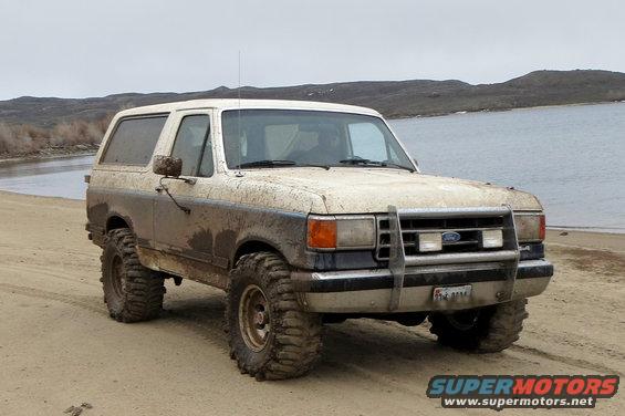 Mud-splattered white Ford Bronco with aftermarket grille guard and large tires parked on a sandy beach