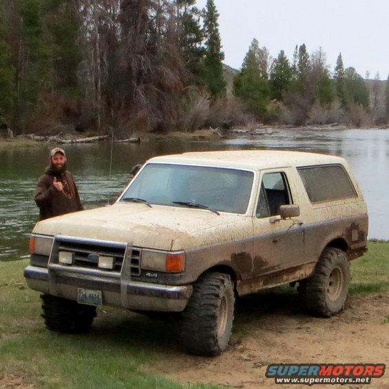 Muddy two-tone Ford Bronco parked by a river with a guy giving a thumbs up