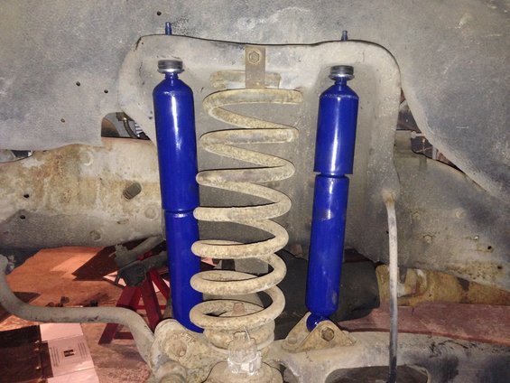 Front suspension components: two blue shock absorbers and a large coil spring. Rusty metal frame visible