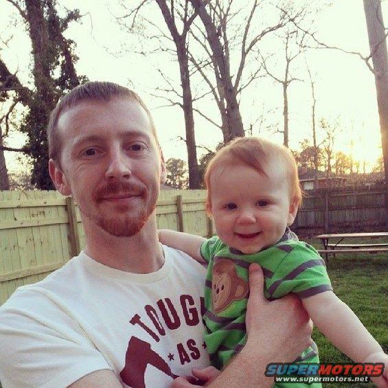 Man holding a smiling baby in a backyard. Man wears a white t-shirt with text. Baby wears a green striped shirt