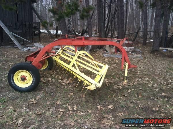 Red and yellow farm hay rake with yellow wheels, parked outdoors near a wooden structure