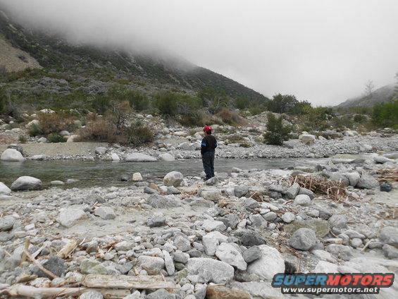 Person in red hat stands in a rocky riverbed with mountains in the background under a foggy sky