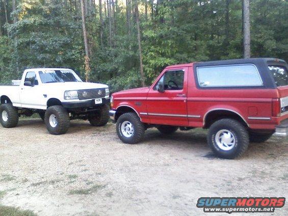 Red 1992 Ford Bronco with black top, lifted on large tires, parked next to a white lifted Ford F-150 truck