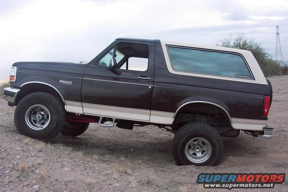Side view of a dark brown and tan Ford Bronco with a white top. It's parked on dirt with a flat tire on the rear passenger side