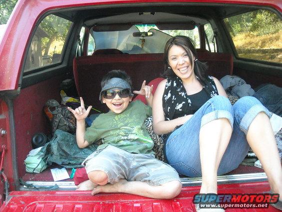 Kid and woman sitting in the open tailgate of a red SUV, surrounded by camping gear