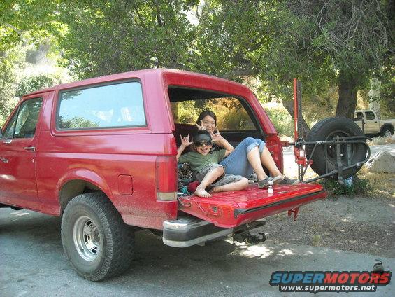 Red Ford Bronco with tailgate open, two people sitting inside. Spare tire mounted on rear. Another truck visible in background