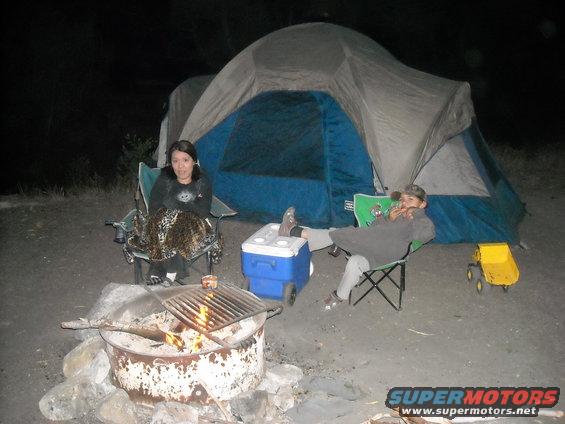 Two people sit in camping chairs by a fire pit and tent at night. A cooler and toy dump truck are nearby