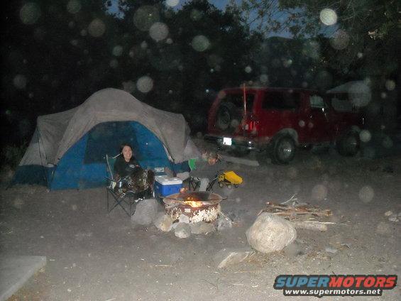 red 1988 Ford Bronco parked next to a tent at a campsite with a campfire going