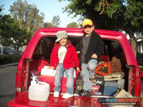 Two kids sit in the open tailgate of a red pickup truck loaded with camping gear