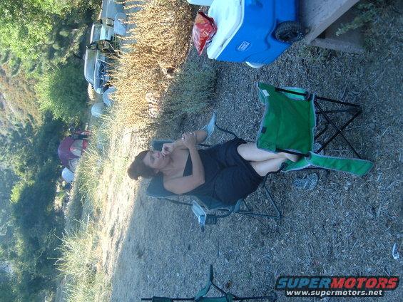 Woman in a black dress sits in a camping chair, with tents and vehicles in the background