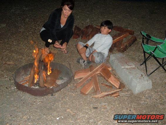 Campfire scene with two people, firewood, and camping chairs