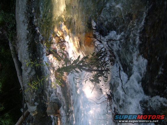 Waterfall flowing over rocks with branches in foreground. Watermark reads SUPERMOTORS