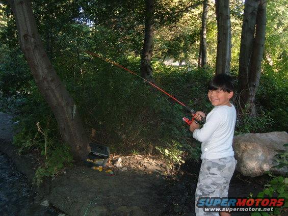 Boy holding a fishing rod and smiling, standing near trees and a stream. A tackle box is visible on the bank