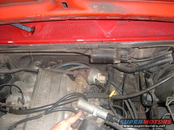Close-up of a Ford engine bay with various hoses and components visible. A hand is pointing to a part