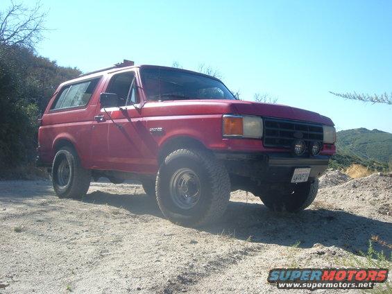 Red Ford Bronco parked on a dirt road, front 3/4 view. Aftermarket wheels and lights are visible