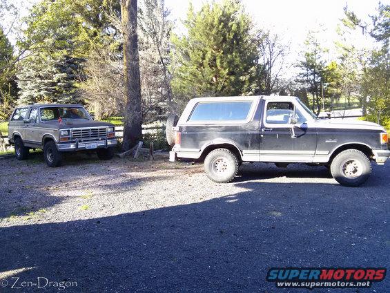 Two Ford Broncos parked on gravel. One is black, the other is brown and grey. Trees and a fence are in the background