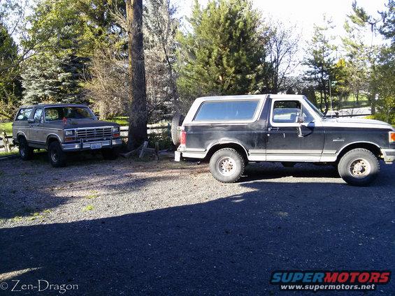 Two Ford Broncos parked on gravel. One is dark with a white top, the other is tan and parked further back