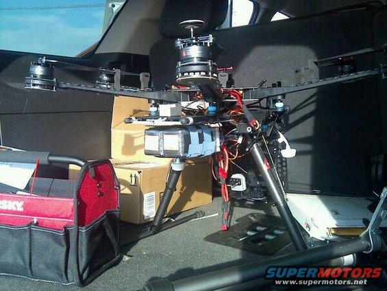 Large drone with multiple rotors and camera mount sits in the back of a vehicle next to a toolbox and cardboard boxes