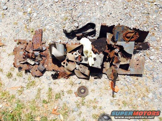 Pile of rusted metal scraps and seatbelts on gravel. A pair of white gloves is on top