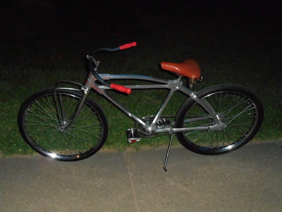 Chrome cruiser bicycle with red grips and a brown seat, parked on a sidewalk at night