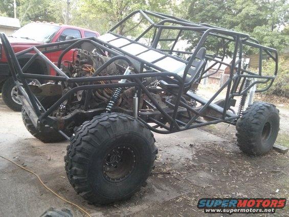 Custom buggy with roll cage, exposed engine, and large tires, parked next to a red pickup truck