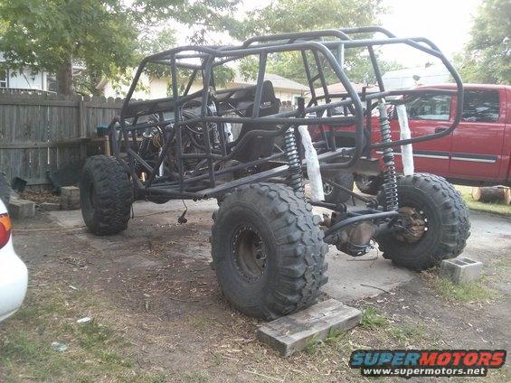 Off-road buggy frame with large tires and suspension components, parked outdoors next to a red pickup truck