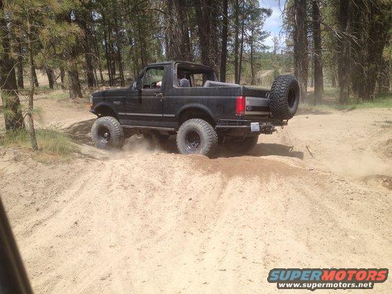 Black Ford Bronco with a spare tire mounted on the rear, driving on a sandy trail with trees in the background