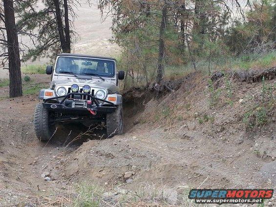 Silver Jeep Wrangler driving off-road on a dirt path, front bumper with winch, trees and dry brush in background
