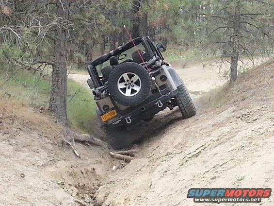 Tan Jeep Wrangler on a dirt trail, rear-left quarter view, with spare tire mounted on the back