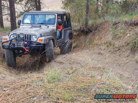 Silver Jeep Wrangler Rubicon driving on a dirt trail with trees around. It has a winch and off-road lights