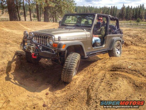 Driver in a tan Jeep Wrangler Rubicon on a dirt trail. The vehicle has a winch and off-road tires