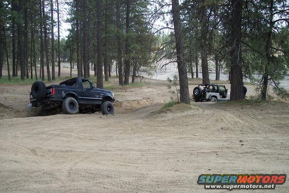 Black Ford Bronco and white Jeep Wrangler driving on a dirt path in a forest