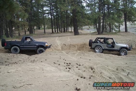 Black 1990 Ford Bronco driving on a sandy trail next to a grey Jeep Wrangler