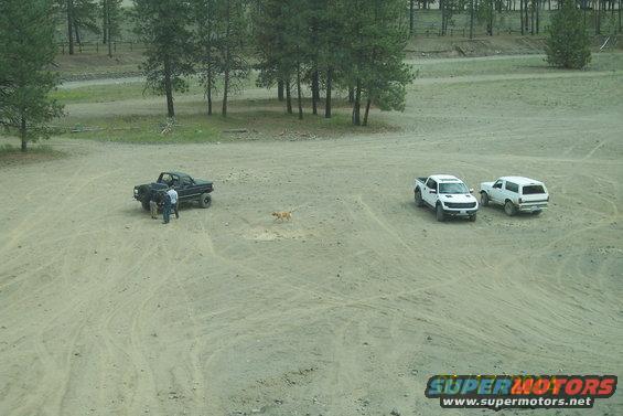 Black pickup truck with people nearby, white pickup truck, and white SUV parked in a dirt lot with trees in background