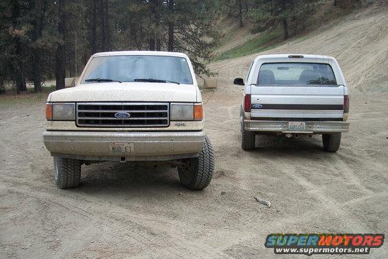 Two Ford Broncos parked on a dirt road, one facing front, the other facing rear. The front Bronco is a 1987 model