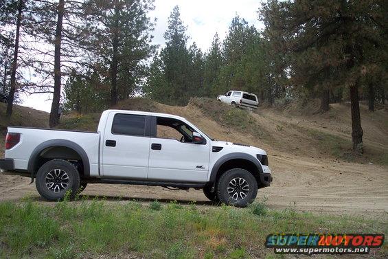 White pickup truck in foreground, white SUV climbing dirt hill in background