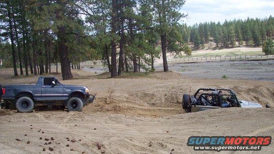 Dark blue pickup truck and a light-colored off-road buggy in a sand pit with trees in background