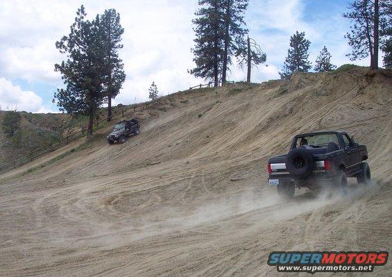 Two off-road vehicles driving up a steep sand dune, one in the foreground kicking up dust