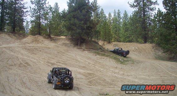 Black Jeep Wrangler with roll cage seen from rear, driving on sand. Pickup truck is visible further up the hill