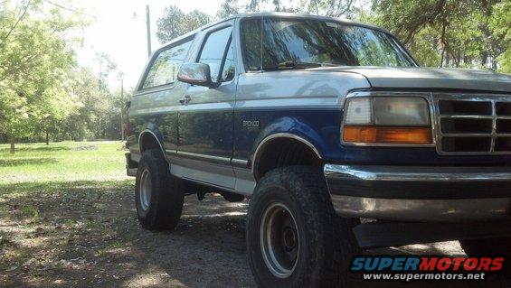 Two-tone blue and silver Ford Bronco parked on a dirt road with trees in the background