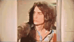 Man with long, wavy brown hair and a denim shirt looks to the side