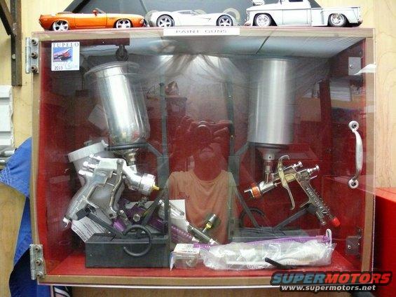 Display case with two paint spray guns, a man's reflection, and model cars on top