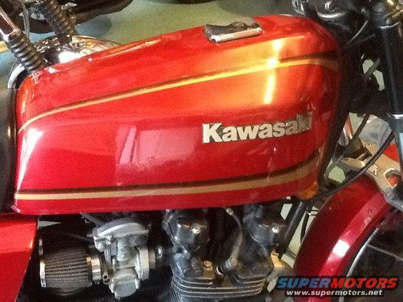 Red motorcycle fuel tank with gold stripes and 'Kawasaki' logo. Engine parts and air filter visible below