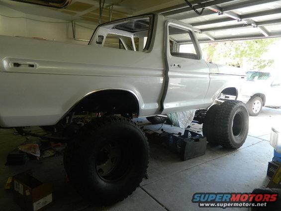 White 1978 Ford Bronco body with large tires, undergoing restoration in a garage. Another white SUV is visible in the background