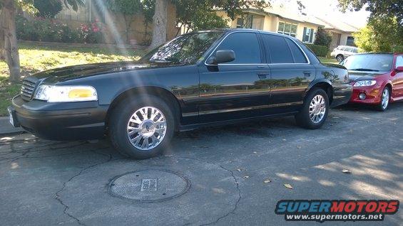 Black 2003 Ford Crown Victoria sedan parked on a street, side view, with a red car behind it