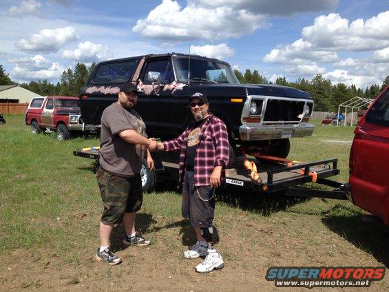 Two guys shaking hands next to a black 1978 Ford Bronco on a trailer. Another red and white Bronco is parked behind it