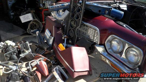 Maroon car front end with grille and headlights, surrounded by disassembled interior and engine parts
